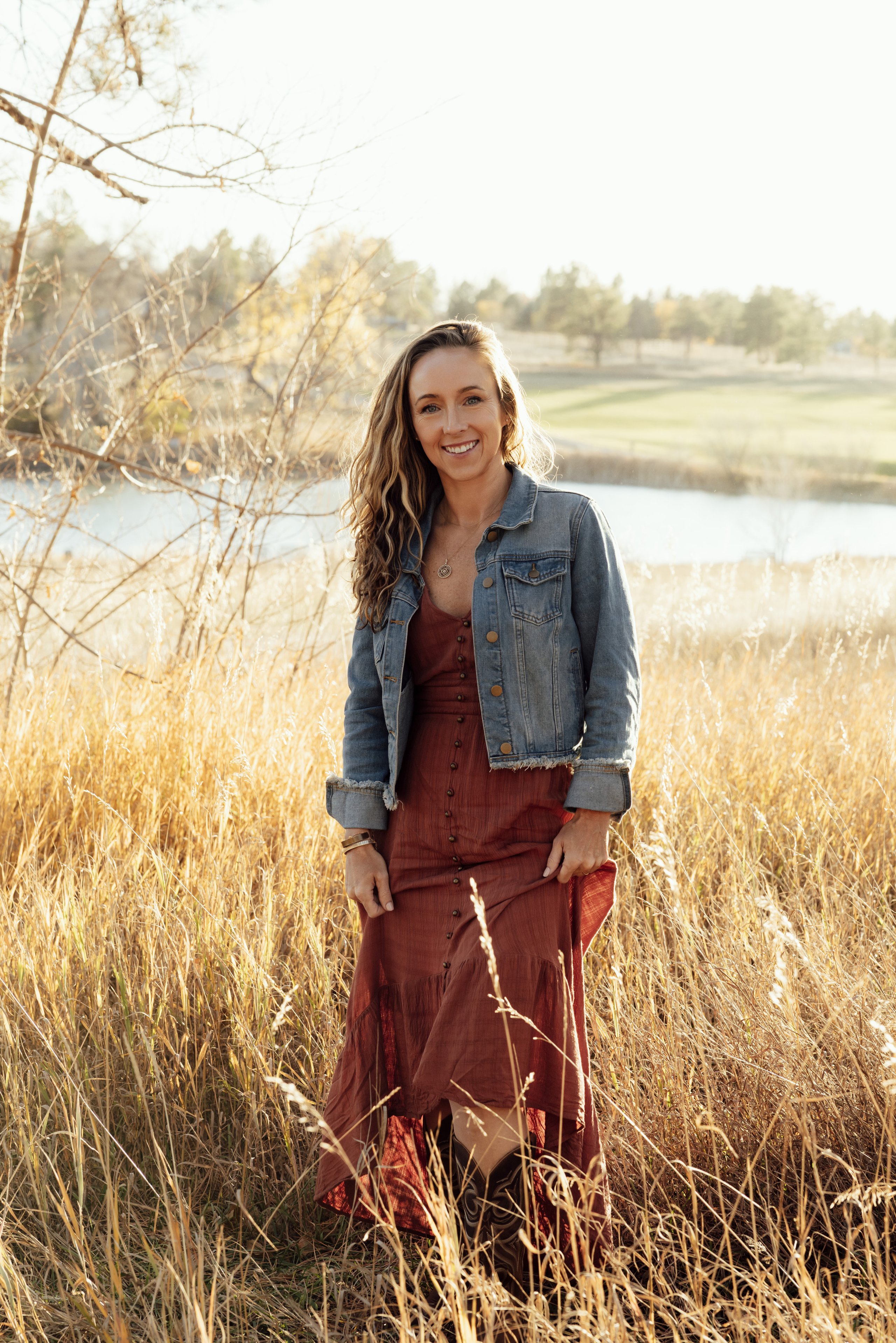 A smiling woman in a denim jacket and rust maxi dress stands in a sunlit field of tall dry grass beside a pond.