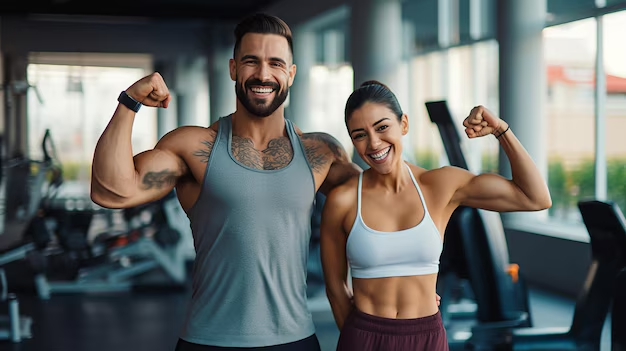 Two smiling gym-goers flex their arms in front of blurred exercise equipment and large windows.