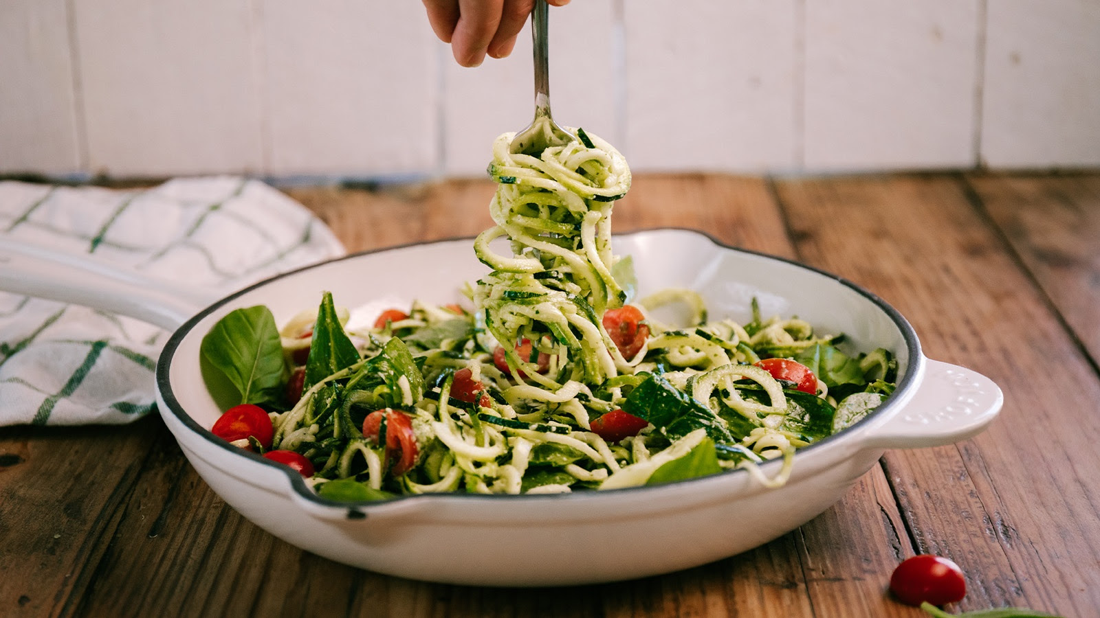 Fork lifting spiralized zucchini noodles from a green salad with cherry tomatoes in a white serving dish.
