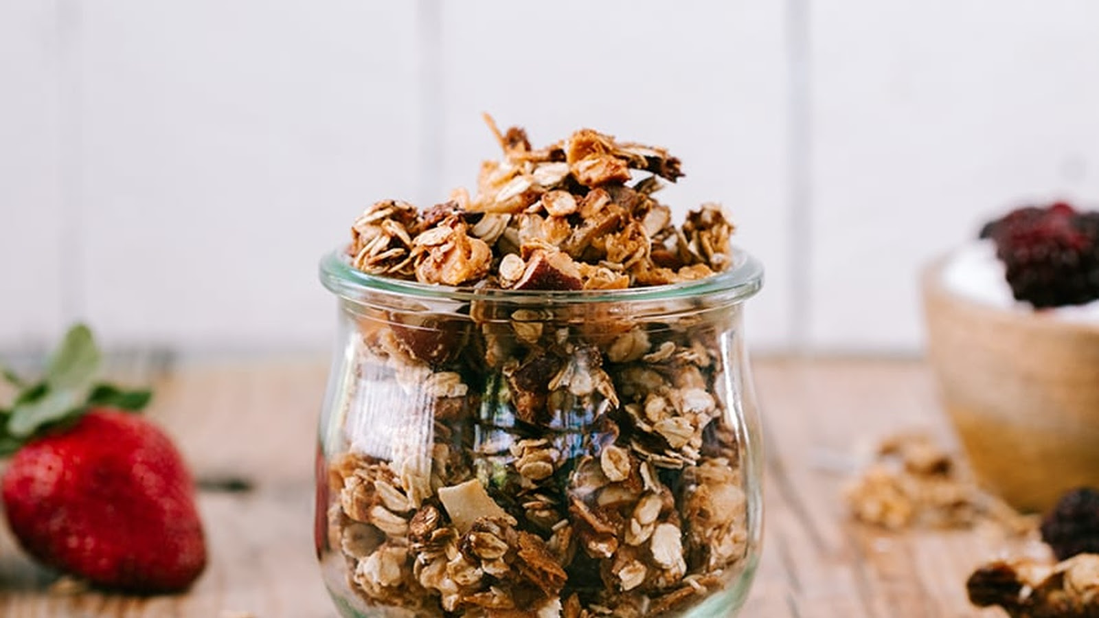 Glass jar overflowing with granola on a table, with a strawberry and blurred berry-topped dish nearby.